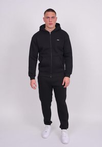 Sweat à capuche noir avec fermeture éclair et pantalon assorti en tissu doux. Comprend deux poches avant et un petit logo sur la poitrine. Porté avec des baskets blanches.
