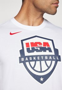 Bílý bavlněný tričko s velkým logem USA Basketball v námořnické modři a červené. Logo Nike v červené na rameni. Kulatý výstřih.