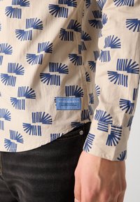 Beige langærmet skjorte med et blåt geometrisk mønster, der har et Scotch & Soda mærke i kanten og en knappet manchet detalje.