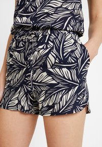 Pantaloni corti blu navy con un motivo a foglia bianca, cintura elastica e tasche laterali. Tessuto leggero con vestibilità comoda e piccole fenditure laterali.