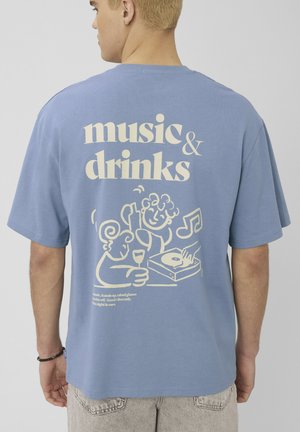 Personne portant un T-shirt bleu avec le texte « musique & boissons » et un dessin animé de deux personnes, une platine et des notes de musique dans le dos.