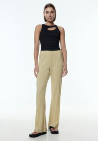 Zwarte mouwloze top met een uitsnijdetail, gecombineerd met hooggesneden, geplooide beige broek en platte zwarte sandalen.