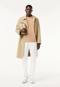 Impermeabile beige sopra una t-shirt color sabbia, pantaloni bianchi e sneakers chiare. Tiene una borsa leggera con un sottile motivo geometrico.