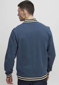 Kék pulóver hosszú ujjakkal, fekete-fehér bordázott nyakkal és mandzsettákkal, sima textúrával és minimális részletezéssel.