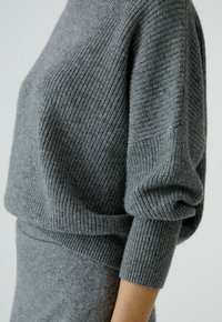 Close-up van een persoon die een grijze geribde gebreide trui met lange mouwen en bijpassende grijze gebreide broek draagt.