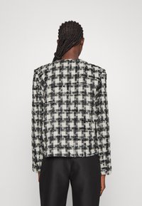 Personne aux cheveux tressés portant une veste texturée à carreaux noirs et blancs et un pantalon noir, vue de dos sur un fond uni.