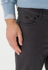 Grijze katoenen broek met een voorzak, subtiele stikdetails en een slanke pasvorm. Hand rustend op de zijkant van de broek.