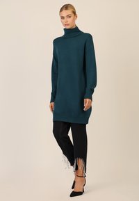 Teal oversized turtleneck sweater med ribbede manchetter og kant, parret med sorte jeans med frynsekanter prydet med perler. Sorte hælede sko.