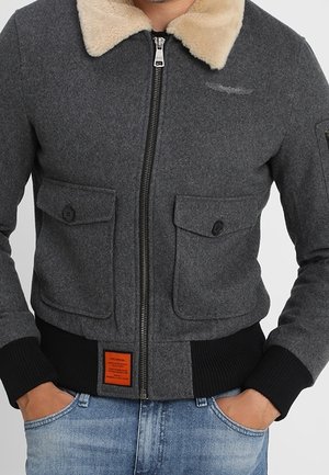 Mand iført en grå jakke med lynlås og beige pelskrave, to forreste lommer med knapper, sorte ribstrikkede manchetter og blå jeans.