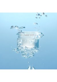 Klarer, quadratischer Glasbehälter mit Creme und einem weißen Deckel, umgeben von Wassertropfen, vor einem hellblauen Hintergrund. Das Etikett trägt die Aufschrift "MAGIC WATER CREAM".