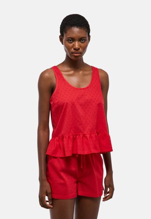 Femme portant un haut rouge texturé sans manches avec un ourlet à volants et un short rouge assorti sur un fond clair uni.