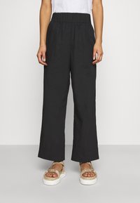 Pantalons larges noirs en tissu léger avec une taille élastique. Associés à des sandales à plateforme beiges avec un design de lanière froncée.