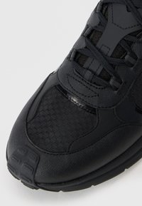 Black sports shoe featuring textured fabric with hexagonal patterns, smooth synthetic accents, and a padded tongue with a lace-up design.
