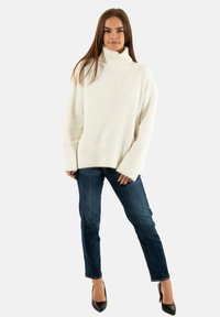 Maglione bianco a collo alto lavorato a maglia, con una vestibilità rilassata, caratterizzato da maniche lunghe e polsini a coste, abbinato a jeans slim-fit blu scuro e tacchi neri.