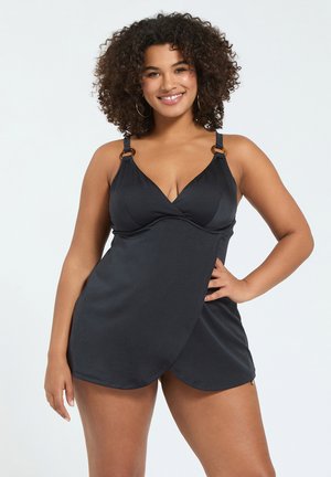 Kobieta o kręconych włosach ubrana w czarną, bezrękawnikową sukienkę kąpielową w stylu kopertowym z regulowanymi ramiączkami, uśmiechająca się, trzymając jedną rękę na biodrze.