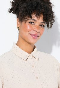 Camisa color beige pálido con cuello tipo camisa, con patrones de puntos plateados por todo ella y cierre de botones en la parte delantera. Pendientes de perlas y oro.