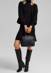 Svart fuskpälskofta över en svart ribbad klänning, i kombination med knähöga svarta stövlar. En strukturerad svart handväska hålls i ena handen.