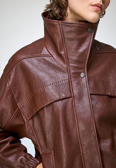 Femme portant une veste en cuir marron avec un col haut et des boutons-pression, cadrée de près pour montrer le haut du torse et le visage partiel.