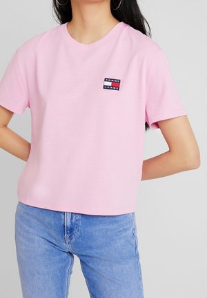 Kobieta ubrana w jasnoróżowy t-shirt Tommy Jeans z krótkimi rękawami i niebieskie dżinsy, stoi z rękami za plecami.