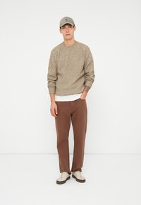 Beige knit sweater, brown pants, and sneakers. Model stands against a white background, wearing a gray cap. Cozy texture, casual fit.