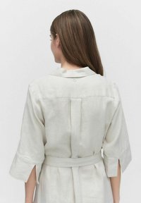 Jasno beżowa lniana sukienka z kołnierzykiem, krótkimi rękawami i paskiem do przewiązania w talii. Posiada detal w postaci plisowania z tyłu.