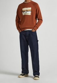 Persona che indossa un felpa color ruggine con il logo "Pepe Jeans London", jeans blu scuro e sneakers nere basse con suola bianca.
