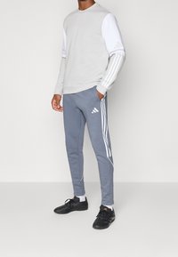Sweat-shirt gris à manches blanches, avec trois bandes blanches. Pantalon de survêtement gris avec trois bandes, coupe ajustée, et chaussures de sport noires.
