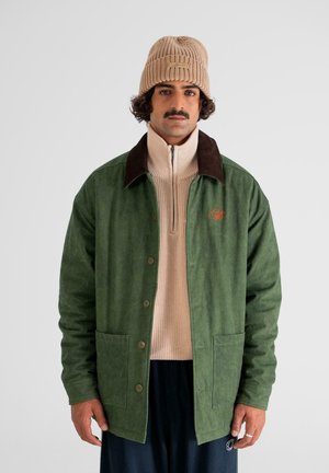 Green cotton jacket with brown collar, front button closure, and two front pockets. Worn over a beige knitted sweater. Brown knitted beanie.