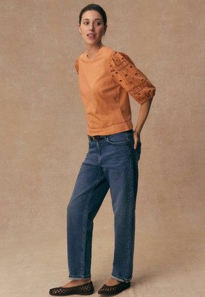 Woman wearing orange puff sleeve top with eyelet details, blue jeans, and black woven flats, standing against a plain beige background.