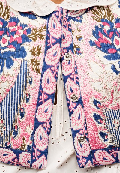 Gros plan sur une veste éclatante à motifs floraux et géométriques en bleu, rose et marron portée sur un chemisier blanc en broderie anglaise.