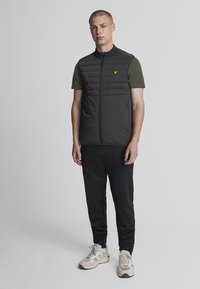 Gilet imbottito nero con dettagli in cuciture verticali, chiusura a zip e un piccolo logo giallo. Indossato su una camicia verde, abbinato a pantaloni neri e sneakers.