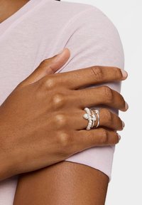 Ręka z trzema srebrnymi pierścieniami z gwiazdkami i perłami spoczywająca na górnej części ramienia, ubrana w jasnofioletową koszulkę z krótkim rękawem.