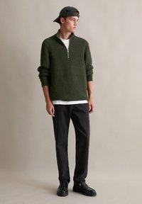 Dunkelgrüner, strukturierter Viertelreißverschluss-Pullover, über einem weißen Hemd getragen, kombiniert mit schwarzen Hosen und schwarzen Schuhen; Modell trägt eine schwarze Mütze.