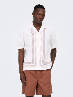 Mann trägt weißes kurzärmeliges Hemd mit vertikaler brauner Stickerei und rostfarbene Cargo-Shorts, steht vor einfachem Hintergrund.