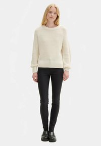 Storas kreminis megztinis su tekstūruotu audiniu, apsiuvtais rankogaliais ir apačia, derinamas su lieknėjančiomis juodomis kelnėmis ir storomis juodomis batais.