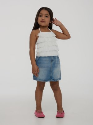 Młoda dziewczyna z długimi ciemnymi włosami, ubrana w biały marszczony top na szyję, jeansową spódnicę i różowe buty, stoi na jednolitym tle.