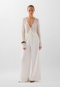 Pulover alb din tricot deschis, cu decolteu adânc în V și talie cu șnur, asociat cu pantaloni largi albaștri. Textură netedă și siluetă lejeră.