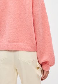 Ροζ φουντωτό πουλόβερ με χαμηλά ώμους, διαθέτοντας ριμπ μανσέτες και στρίφωμα. Κρέμ παντελόνι με λεπτομέρεια φιόγκος στην πλευρά.