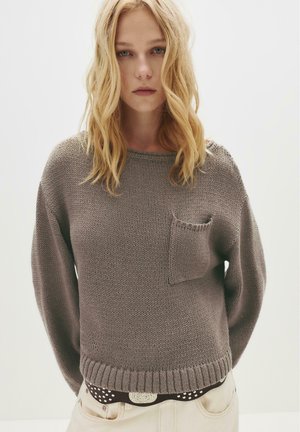 Junge Frau mit blonden Haaren trägt einen braunen Strickpullover mit Brusttasche über einem braun gemusterten Hemd, kombiniert mit hellen Hosen und einem Gürtel.