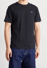 Černé tričko s krátkým rukávem z bavlny, s malým modrým logem na hrudi a volným střihem. Nosené s modrými džíny.