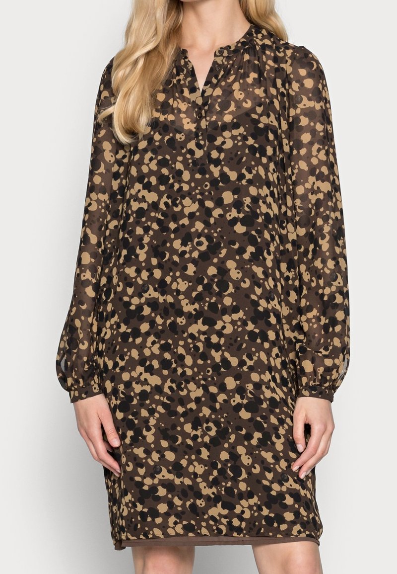Femme portant une robe ample à manches longues, longueur genou, avec un motif tacheté marron et noir et un petit col en V.