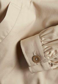 Beige bluse detaljeret med samlet manchet og en enkelt knap. Stoffet viser fin tekstur og syninger langs kanterne.