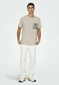 Camiseta de manga corta beige con un bolsillo estampado de hojas, combinada con pantalones blancos y zapatillas crema. Tejido suave, corte relajado.