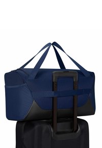 Τσάντα duffel σκούρου μπλε χρώματος ασφαλισμένη στη επεκτεινόμενη λαβή μιας μαύρης βαλίτσας με ρόδες, σε λευκό φόντο.