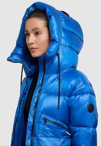 Modrá nadýchaná bunda s veľkou kapucňou, zapínaním na zips vpredu a bočným vreckom. Má lesklý vzhľad a čierne šnúrky na nastavovanie.