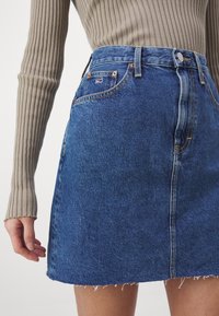 Mörkblå denimminikjol med rå fåll, med framfickor, kn äppning och gula sömnadsdetaljer. Texturerad ribbad topp synlig.