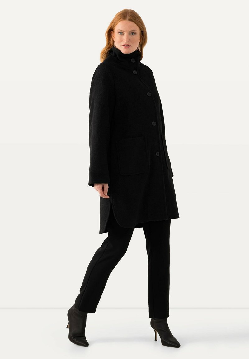 Cappotto di lana nero con colletto alto, chiusura a cinque bottoni, due tasche frontali e aperture laterali. Indossato sopra pantaloni neri su misura e stivali con tacco.