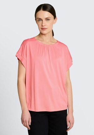 Kobieta o ciemnych włosach związanych nisko w kucyk, ubrana w luźną koraloworóżową bluzkę z krótkim rękawem i czarne spodnie, stojąca na jasnym tle.