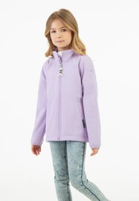 Ung tjej med blont hår som bär en ljuslila zip-up jacka och urtvättade blå jeans, stående mot en vit bakgrund.