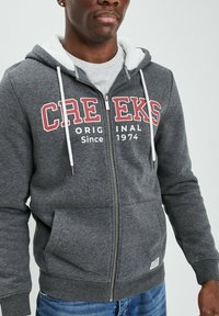 Sudadera gris con cremallera, cordones blancos, bolsillos frontales y forro de felpa. Presenta texto en rojo y blanco: "CREEKS Original Desde 1974."